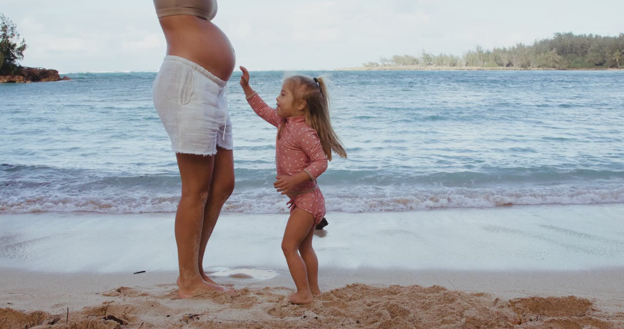 Little Dipper toddler wearing the belly flop crop beachball swimsuit, rubbing a pregnant woman's belly